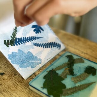 Close-up photo. in focus on the left is a pair of hands pulling an inky leaf fern off of a piece of card covered in blue leaf designs. Beside it on the table top and out of focus is a thick piece of plastic covered with various leaves on top of a faint layer of blue ink.
