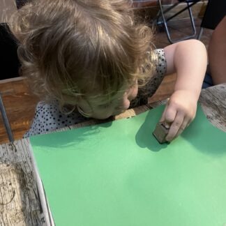 A small child around three years old with blonde hair using a wooden block to stamp onto a piece of green paper