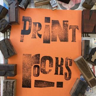 An piece of orange paper with print rocks printed onto it in blacks. The letters are all sizes and fonts and have a wood texture, and the I in print is an upside down exclamation mark. This poser is surrounded by lots of vintage wooden letter blocks scattered around the edges of the paper and the image.