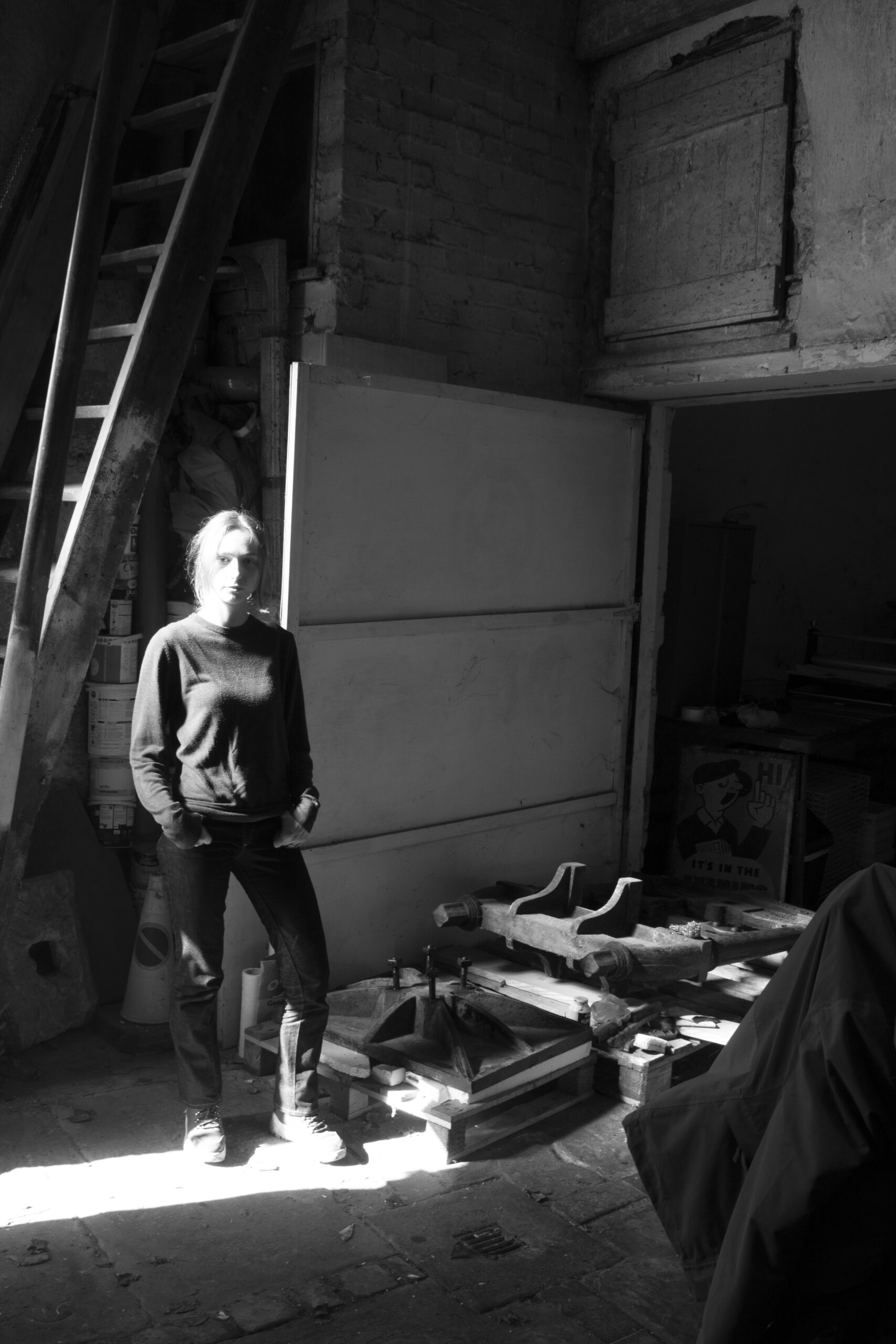 A black and white image of a woman stood in a large cavernous space next to old rusted hunks of metal in front of a large wooden garage door