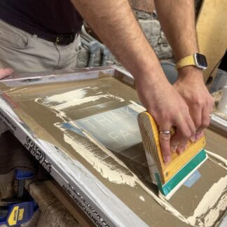 A close-up of someone screen printing in a cream ink. They are holding a squeegee in an upright position and puling it toward them selves.